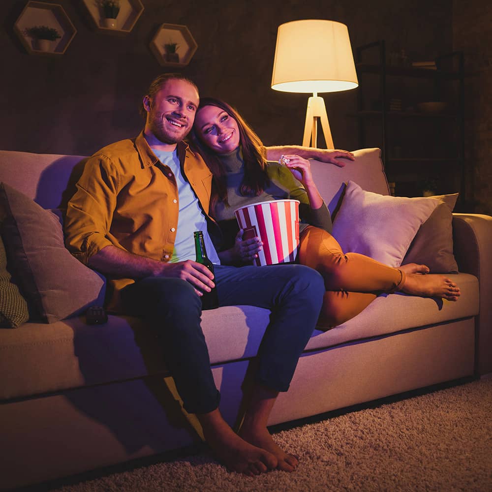 Attractive male/female couple eating popcorn and beer on beige sofa while watching TV.